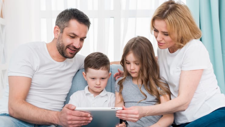 a mother and father sitting on a couch on either side of their two young children. they are all looking at a computer tablet